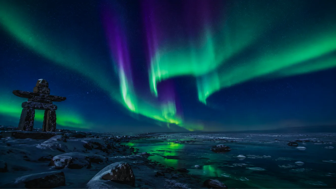 Green and violet northern lights over the shore of Hudson Bay at Churchill, an inukshuk silhouetted at left.