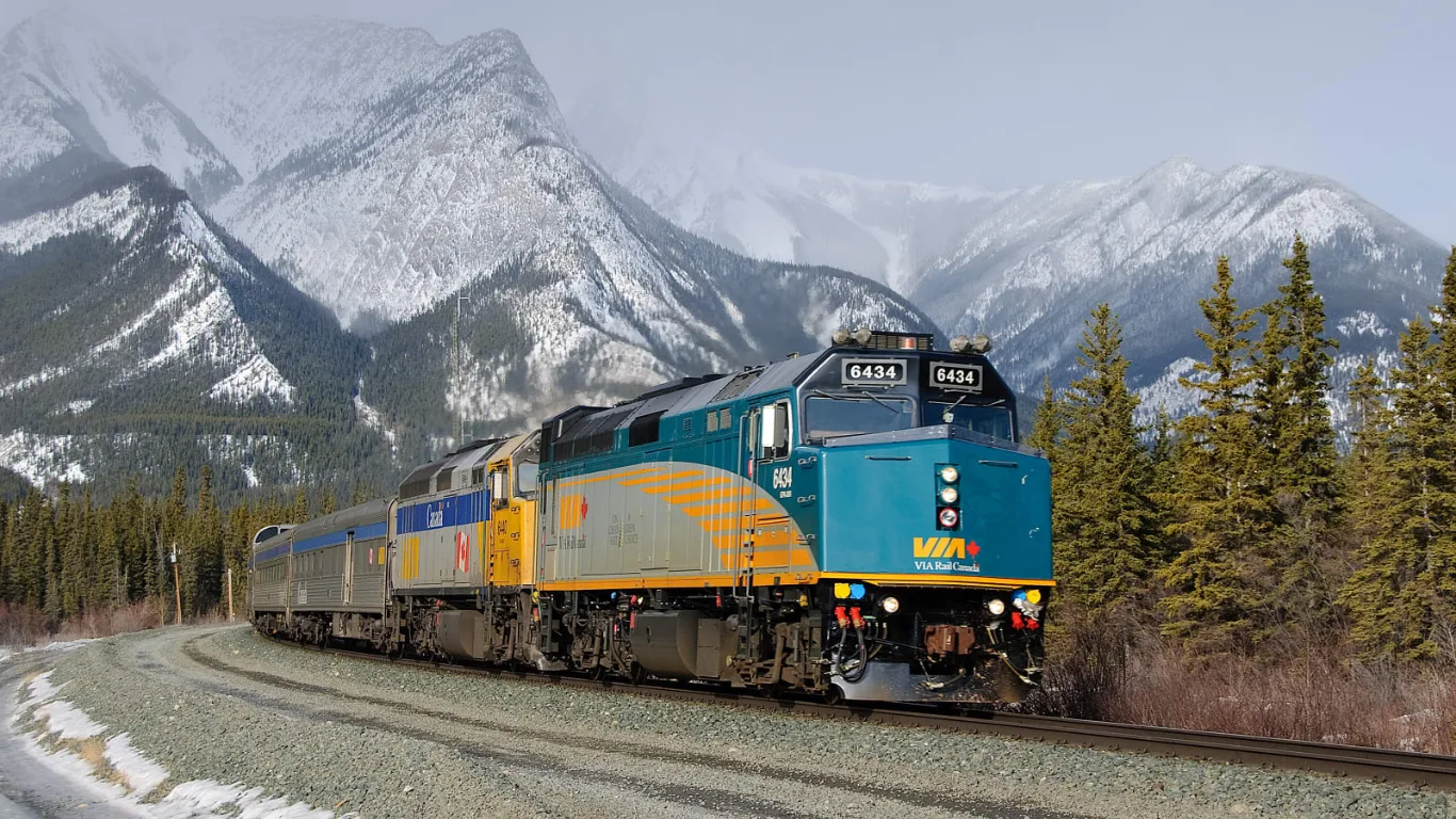 VIA Rail’s The Canadian, led by two locomotives, rounding a curve beneath snow-covered Rocky Mountain peaks.
