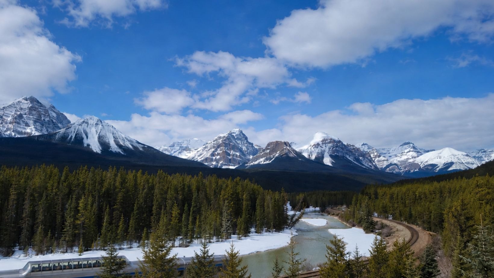 A passenger train curving along a river beneath snow-covered Rocky Mountain peaks under a wide blue sky.